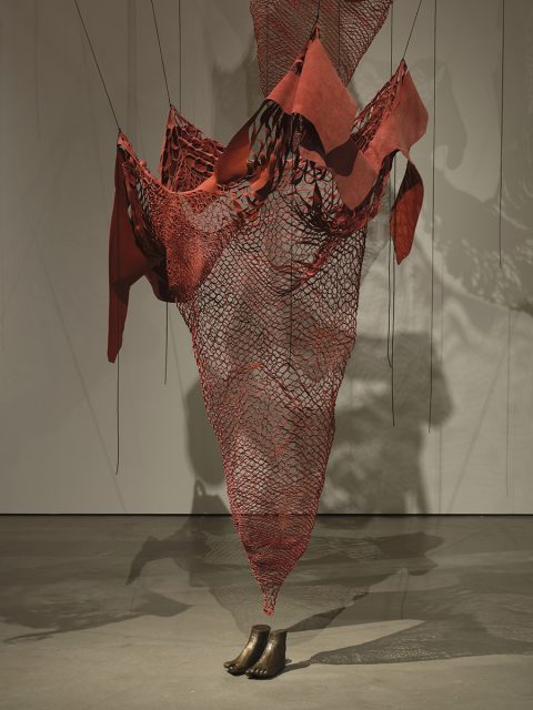 A suspended red mesh and fabric sculpture is anchored by two bronze feet on the floor, with thin cords attaching it to the ceiling and walls.