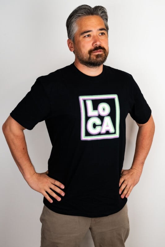 A man with gray hair and a beard stands with hands on hips, wearing a black T-shirt that says “LOCA” in bold, outlined letters. He is looking slightly to the side against a plain white background.