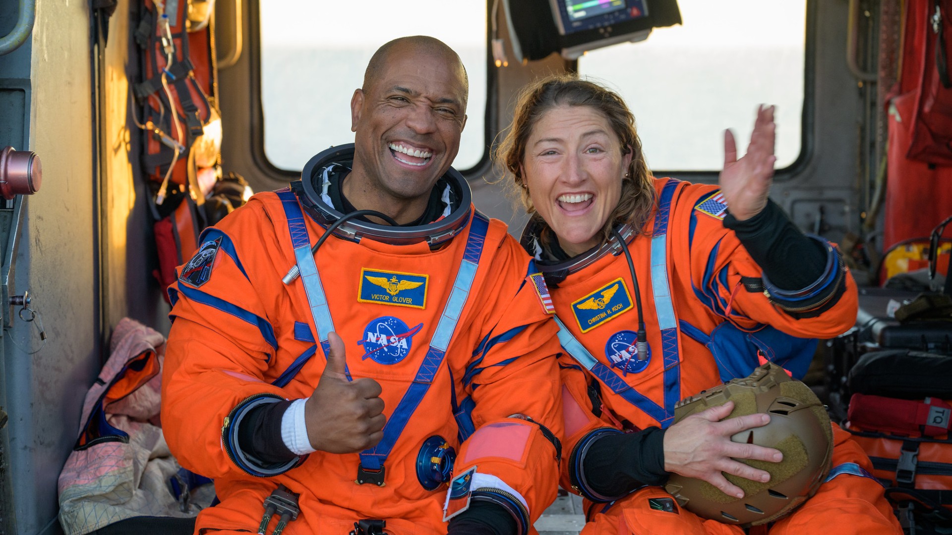 two people in bright orange spacesuits smile and wave inside a helicopter on the deck of a ship at sea