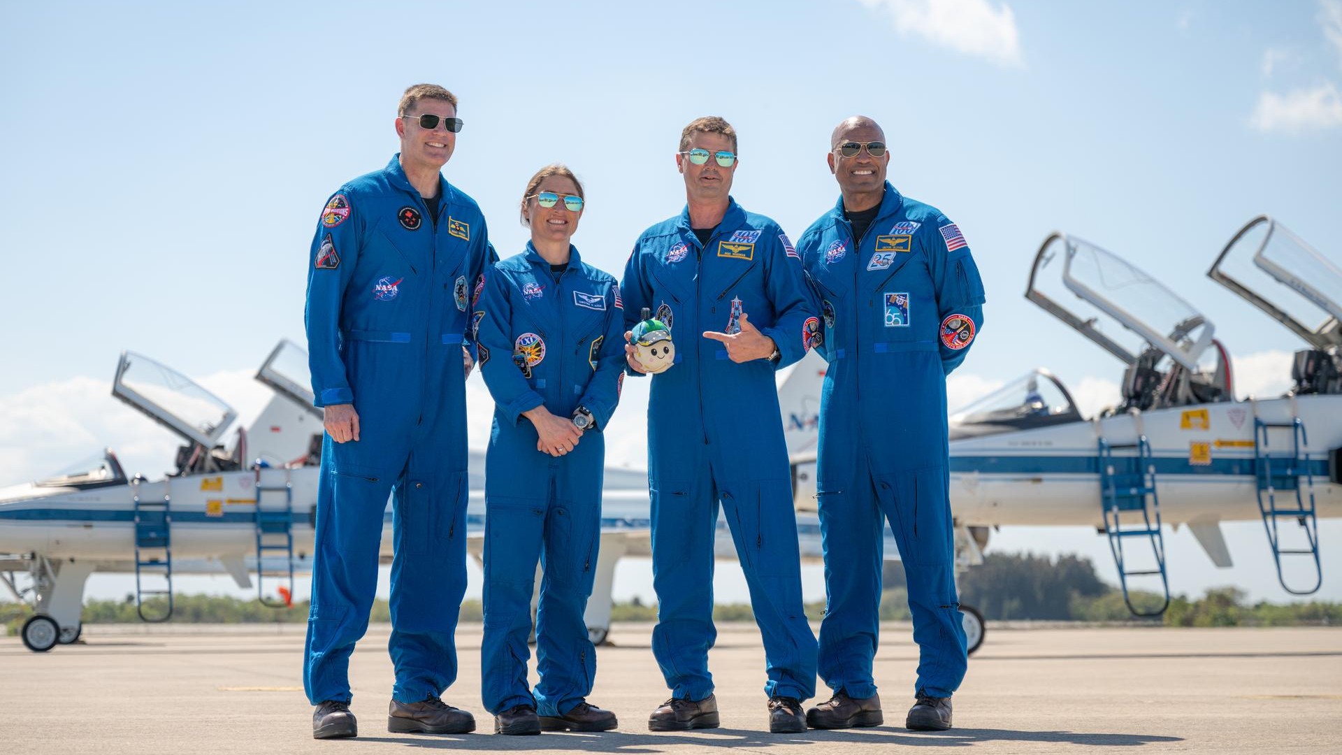 four people in blue flight suits and sunglasses smile for a portrait on an aircraft runway under a sunny sky