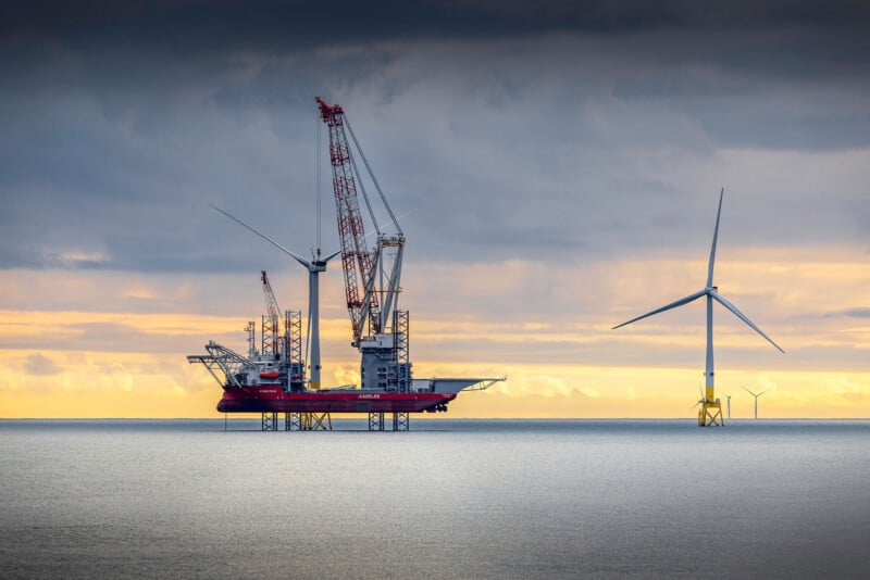 A large offshore construction platform with cranes stands in the sea near wind turbines under a cloudy sky at sunset.