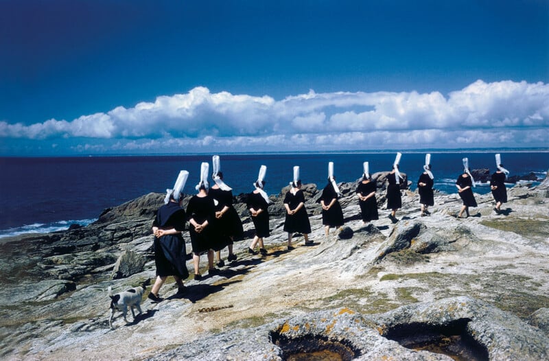 A group of women in black dresses and tall white headdresses walk in a line along a rocky shoreline under a blue sky, followed by a small white dog.