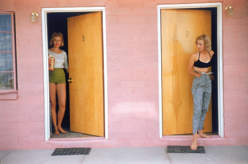 Two women stand in doorways of a pink building. The woman on the left wears green shorts and holds popcorn, while the woman on the right, barefoot, wears plaid pants and a black top, holding a plate. Both doors are slightly open.