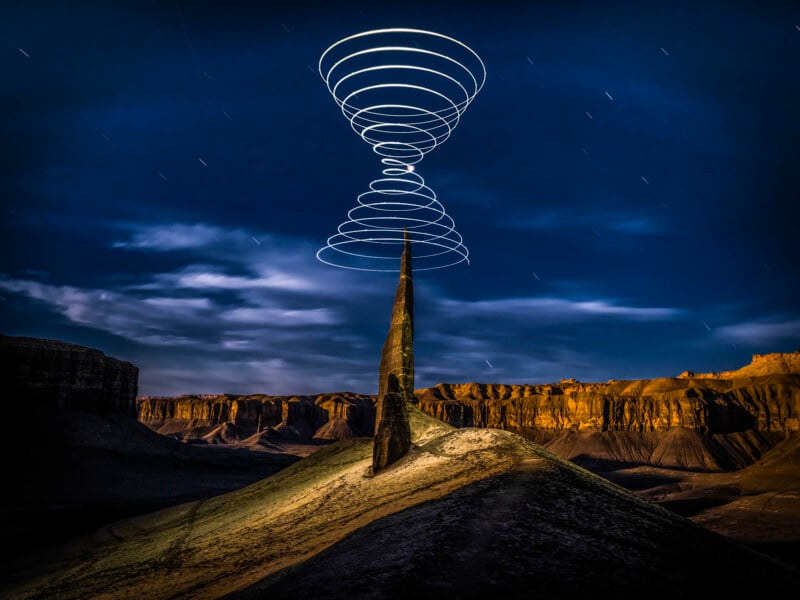 A tall rock spire stands in a desert landscape at night, with light trails forming spirals above and below its peak; distant cliffs are illuminated under a starry sky.
