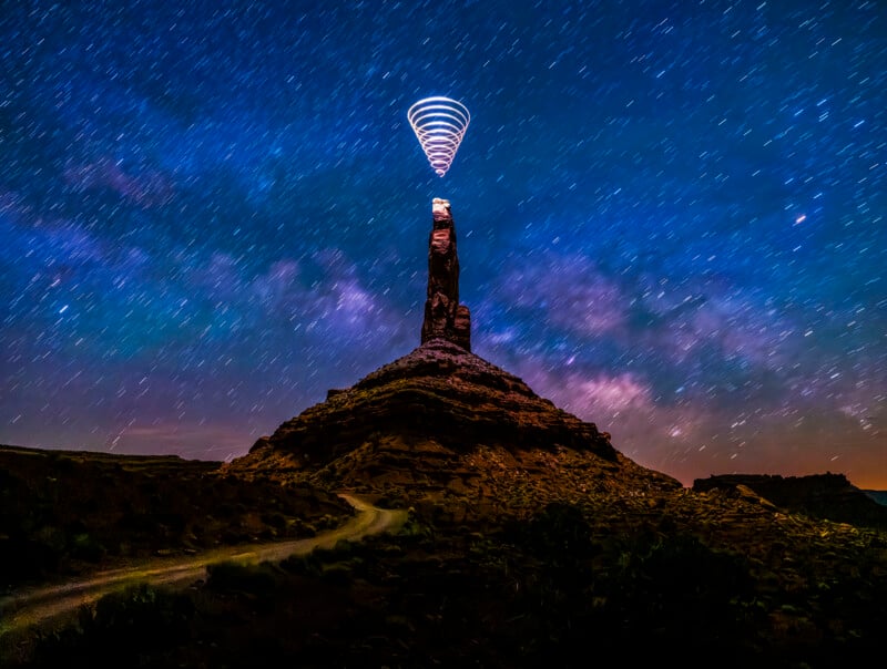 A tall rock formation stands beneath a star-filled night sky, with long-exposure light trails forming a cone shape above its peak, creating a surreal, glowing effect. A winding path leads toward the formation.