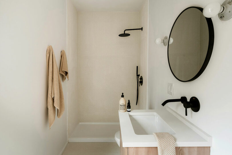 Minimalist bathroom with a walk-in shower, black fixtures, a round mirror, wall-mounted sink, beige towels, and toiletries on the counter.