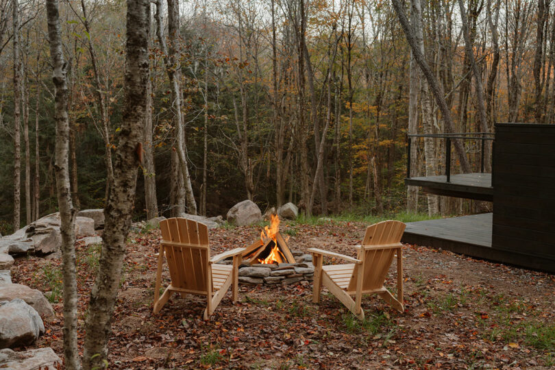 Two wooden chairs face a small stone fire pit with a burning fire, surrounded by trees with autumn foliage and a wooden deck nearby.
