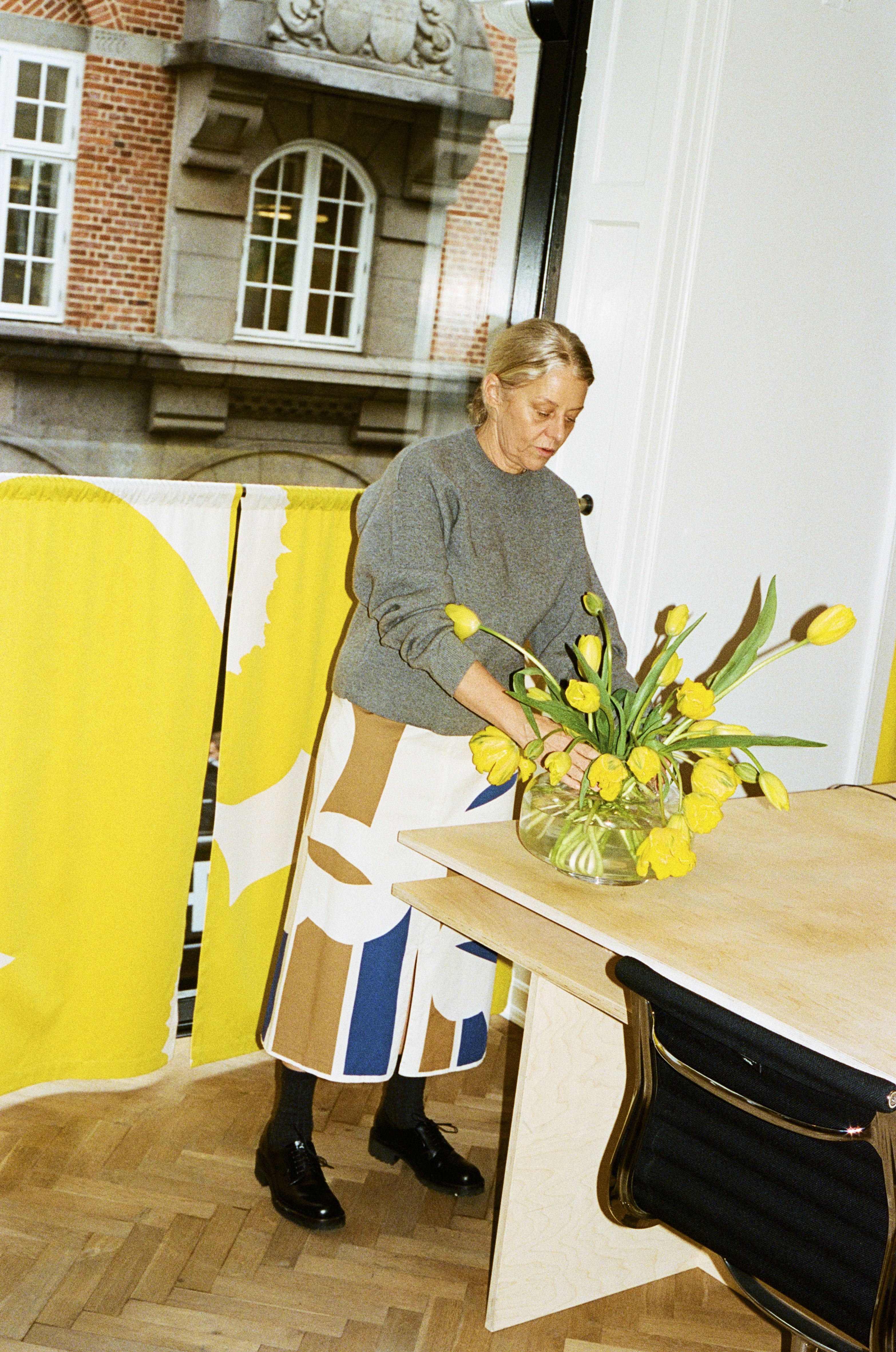 The exterior of an osteria, decorated with a floral mural, a table topped by floral-shaped serveware, and a woman dressed in a gray jumper and a white, brown, and blue skirt, handling yellow tuplis.