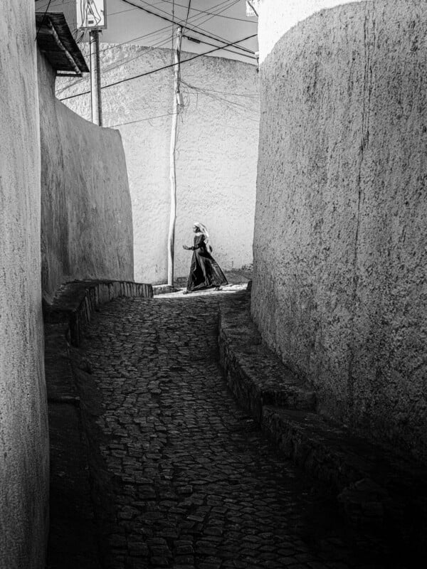 A person wearing a long coat and head covering walks along a narrow, winding cobblestone alley between high, textured walls in a sunlit, deserted street.