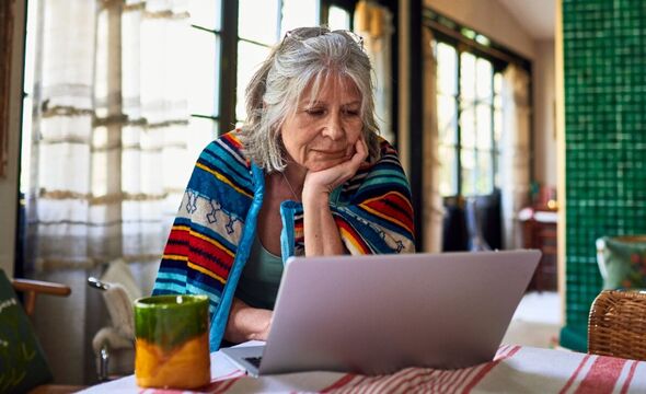 A woman wrapped in a blanket stands at a dining table looking at a laptop