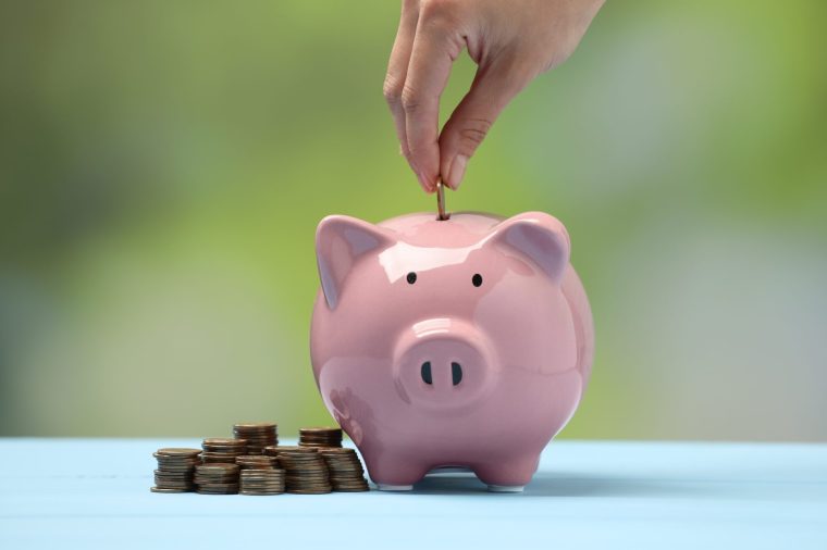 Woman putting coin into pink piggy bank at light blue table against blurred background, closeup - stock photo