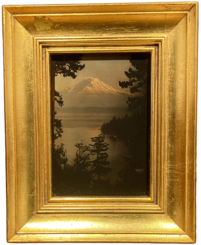 A framed photograph of a snow-capped mountain behind a lake, with pine trees in the foreground. The ornate frame is gold with a distressed finish.