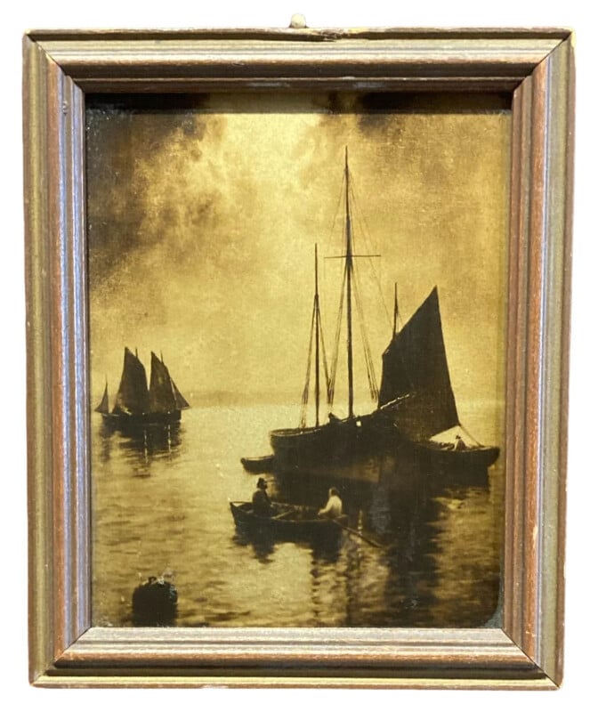 A sepia-toned framed painting shows sailboats and small rowboats on calm water, with figures aboard. The sky appears cloudy, reflecting on the serene surface.