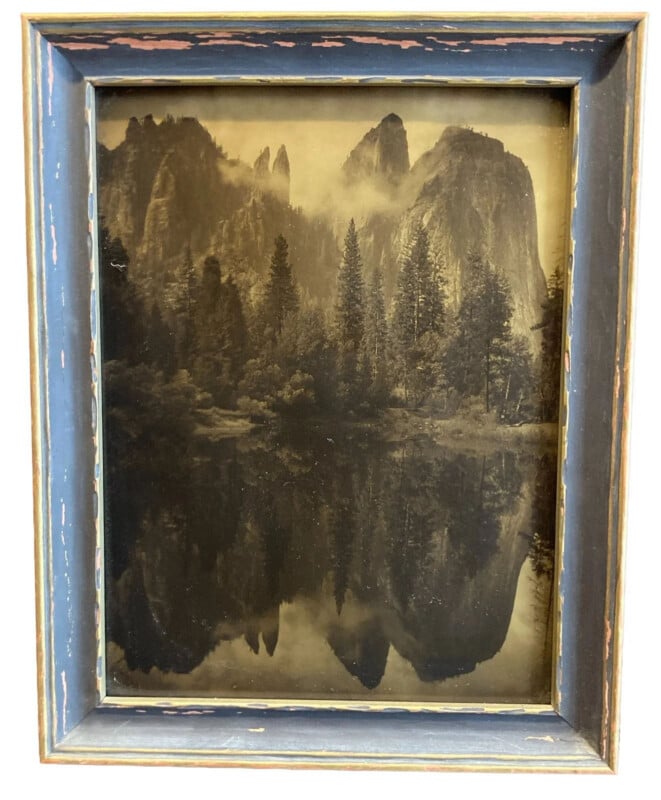 A framed sepia-tone photograph shows tall pine trees and dramatic rocky cliffs reflected in a calm lake, evoking a vintage landscape scene. The frame is rustic with distressed blue paint.