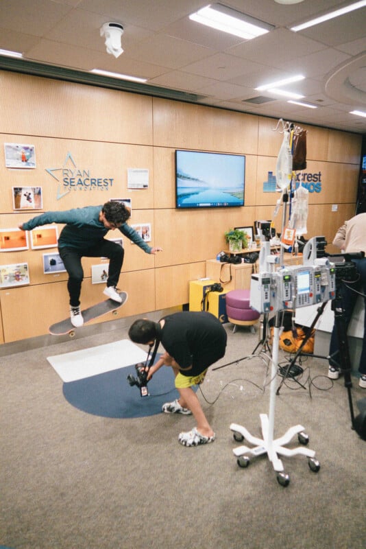 A person on a skateboard does a trick indoors while another person films with a camera. Medical equipment and IV poles are nearby, and the setting appears to be a hospital or medical facility.