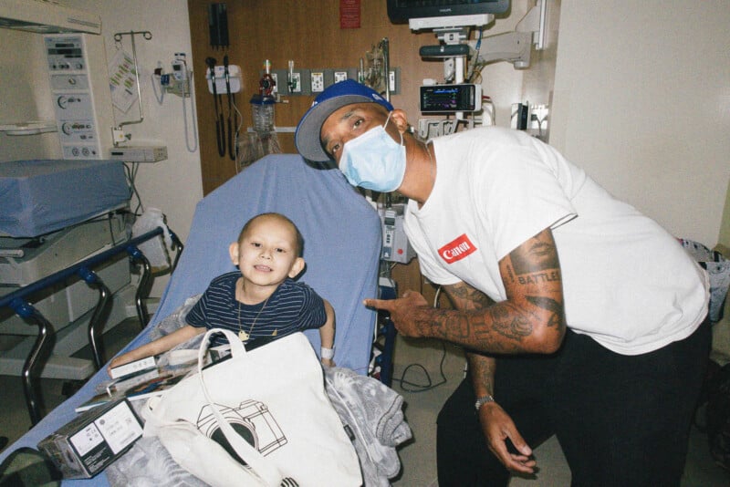 A smiling child sits on a hospital bed, while an adult wearing a face mask and cap kneels beside them, pointing and posing for the photo in a hospital room.
