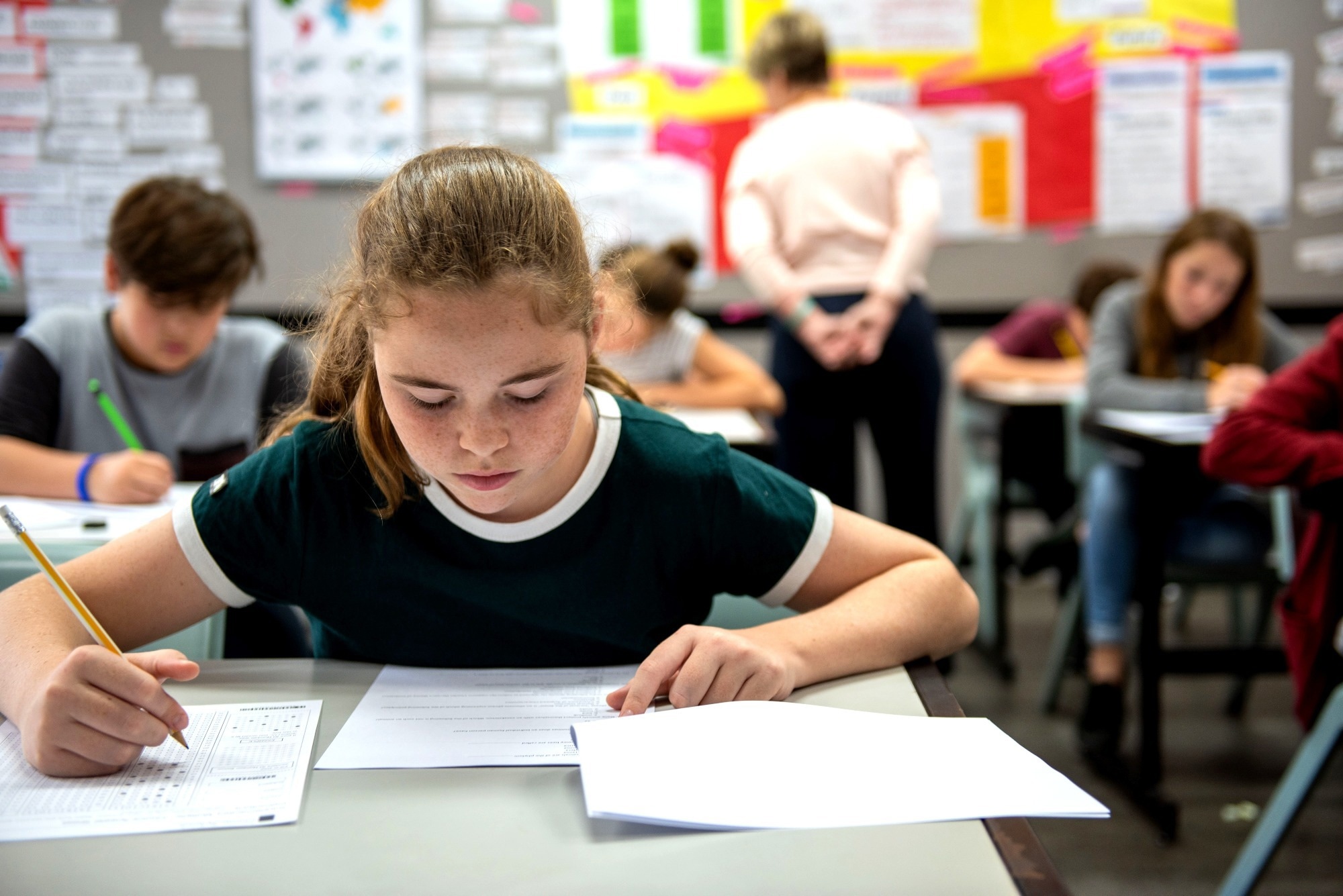 Students in a classroom taking a test, focused on their papers. A teacher observes in the background, ensuring a quiet environment.