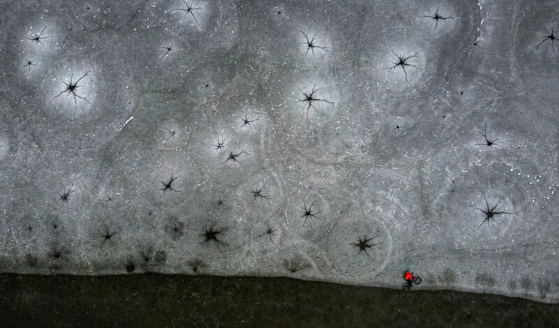 A person in a red jacket rides a bicycle along the edge of a frozen surface covered in dark circular cracks and starburst patterns, seen from above.