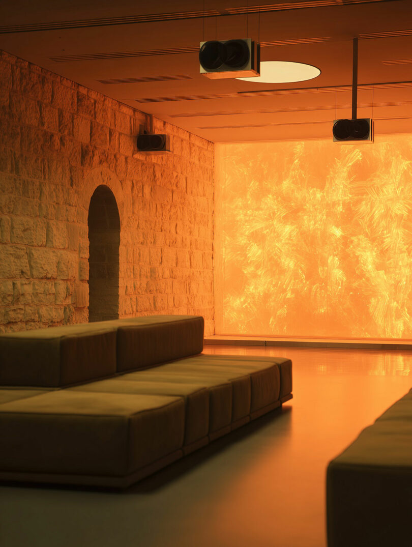 Modern lounge with stone walls, low seating, ceiling speakers, and a large glowing orange backlit wall creating a warm ambiance.