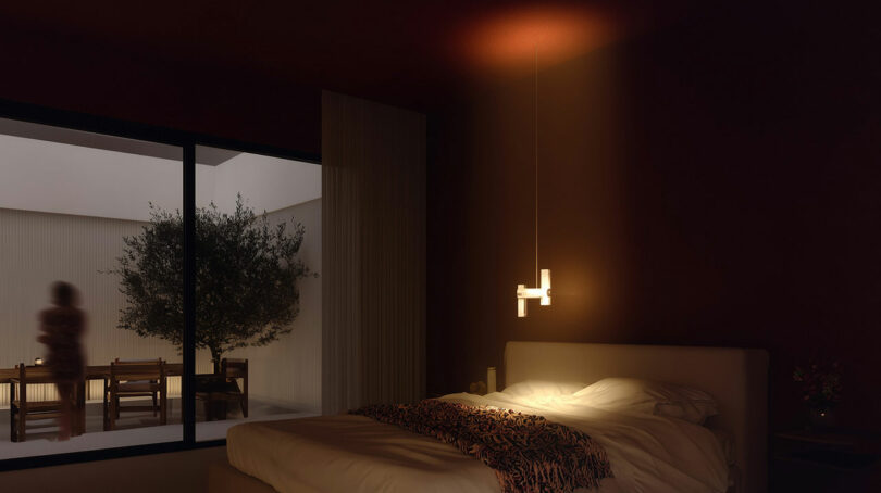 A dimly lit bedroom with an unmade bed, a modern hanging lamp, large window, and a blurred silhouette of a person standing outside near a tree and table.