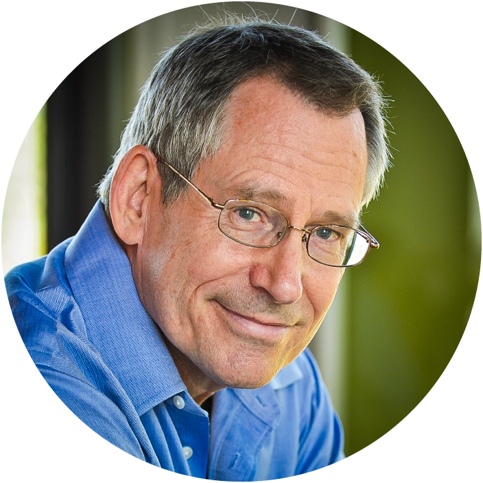 An older man with gray hair and wire-rimmed glasses stands in front of a whiteboard covered in red marker equations and diagrams, holding an orange marker with his arms crossed.