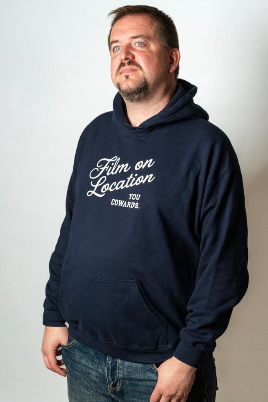A man with short hair and a beard wearing a navy hoodie with the words "Film on Location You Cowards." He is standing against a plain light background, looking slightly upward and to the side.