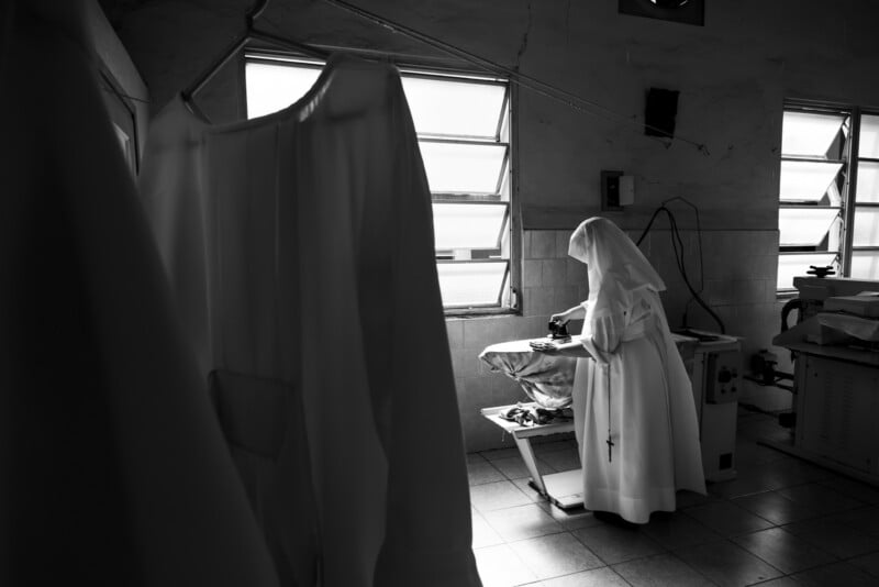 A nun in a white habit stands ironing clothes in a dimly lit room with tiled floors and two windows. A white robe hangs in the foreground, partially obscuring the view.