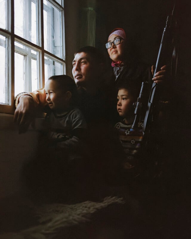 A family of four, including two children, sits closely together by a window, illuminated by daylight. The mother holds a rifle, while all look outside with serious expressions. The room around them is dimly lit.