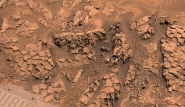A close-up view of a rocky, reddish Martian surface with jagged, layered rock formations and scattered dust. Part of a rover’s equipment marked “CURI” is visible in the bottom left corner.