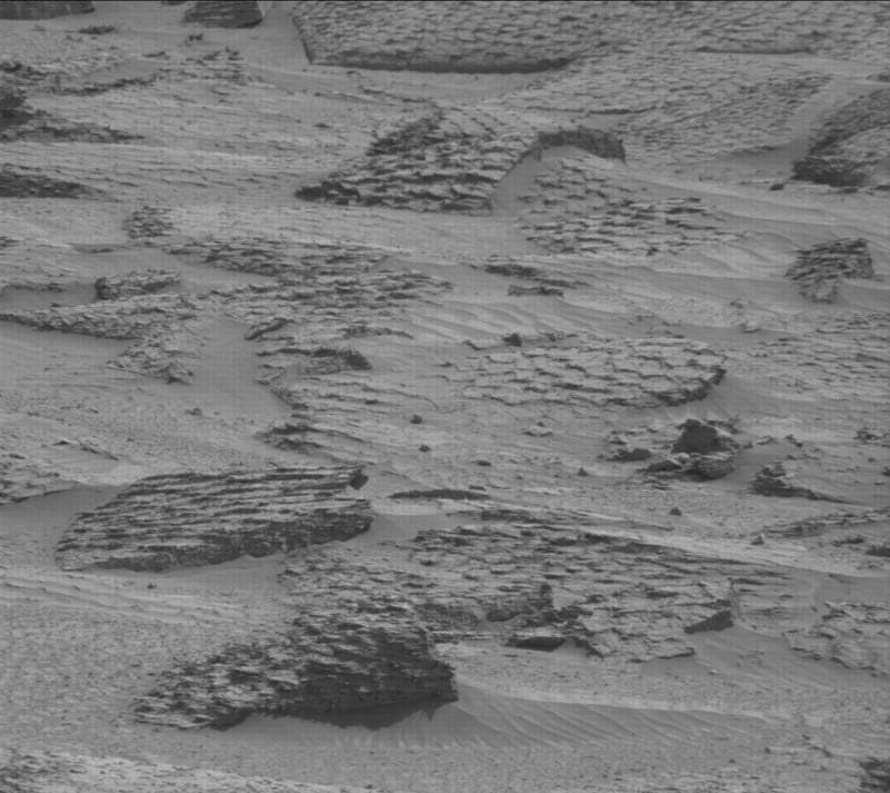 A black and white image shows a rocky, barren landscape with layered rock formations and scattered sand. The terrain appears uneven, with flat, cracked rocks and rippled sand patterns. No vegetation or signs of life are visible.