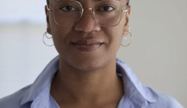 A Black woman with a shaved head wearing gold wire-frame glasses and small hoop earrings in a blue dress shirt.