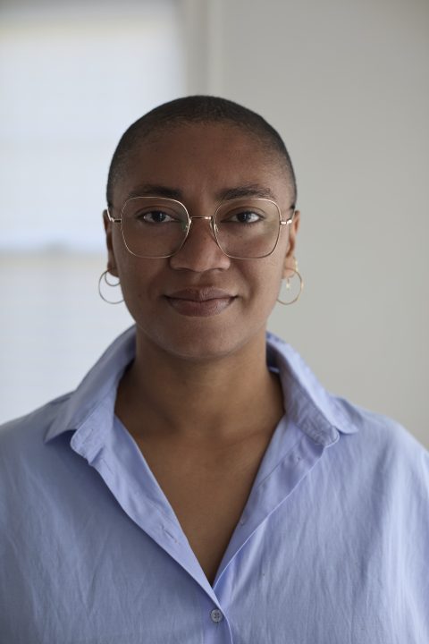 A Black woman with a shaved head wearing gold wire-frame glasses and small hoop earrings in a blue dress shirt.