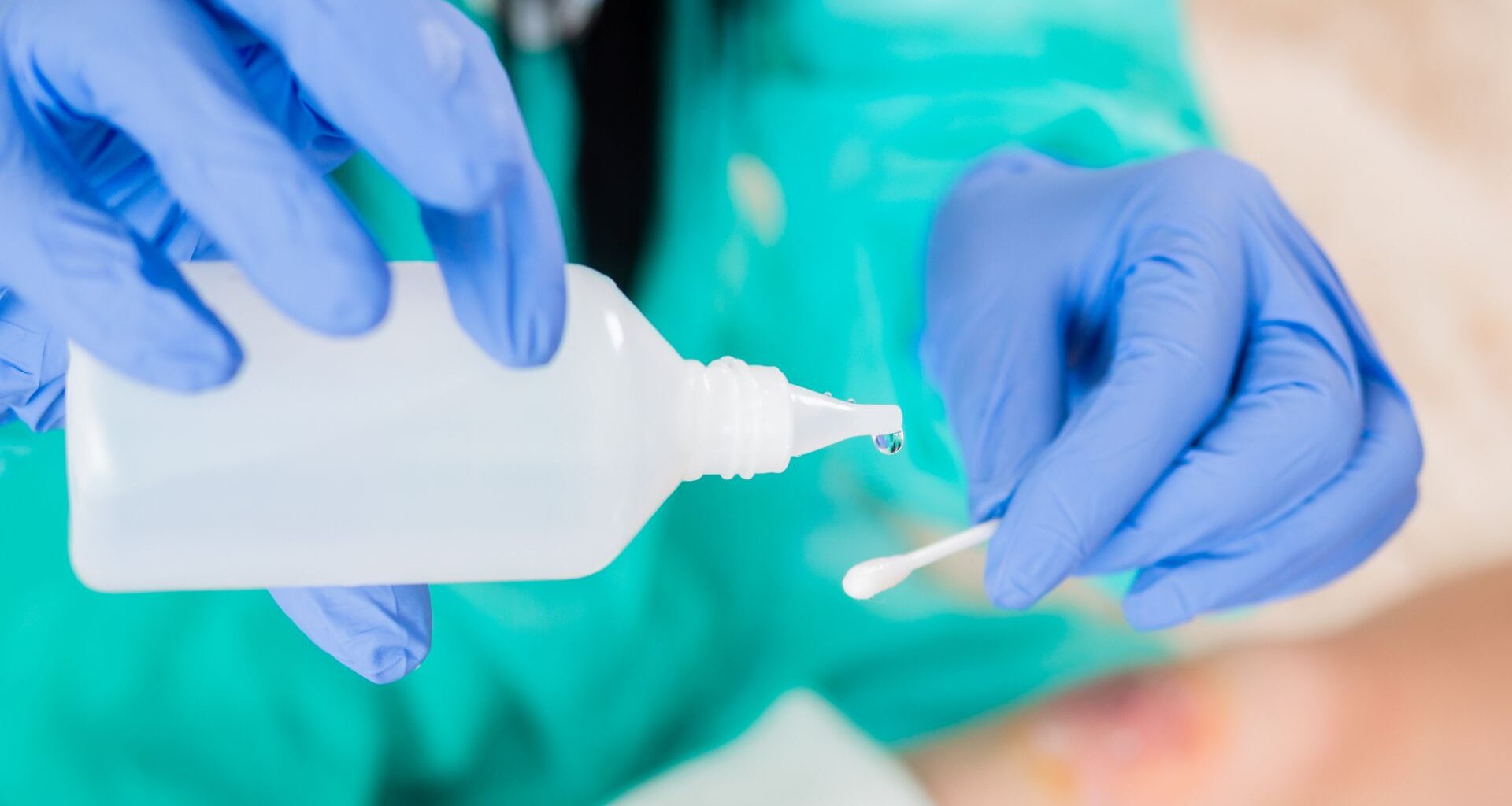 A person wearing turquoise scrubs and blue nitrile gloves pours clear water from a square plastic bottle onto a q-tip