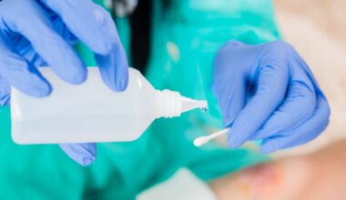 A person wearing turquoise scrubs and blue nitrile gloves pours clear water from a square plastic bottle onto a q-tip