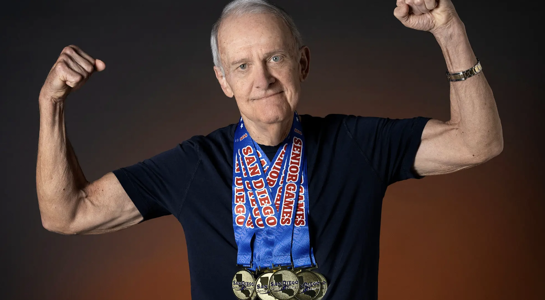 Senior man with arms raised wearing multiple gold medals around his neck, the blue ribbons read &ldquo;San Diego Senior Games&rdquo;