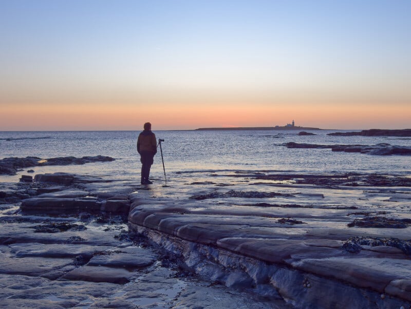 A person with a walking stick stands on rocky coastal terrain, looking out over calm sea water at a distant lighthouse silhouetted against a colorful sunset or sunrise sky.