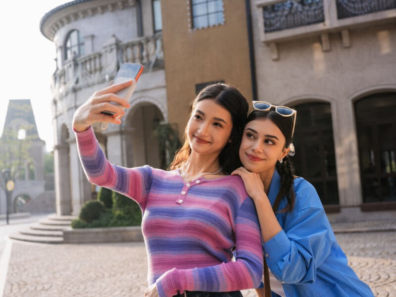 Two young women smile while posing for a selfie outdoors. One, in a pink and purple striped sweater, holds the phone. The other, in a blue shirt with sunglasses on her head, leans on her friend's shoulder. A sunny building is in the background.