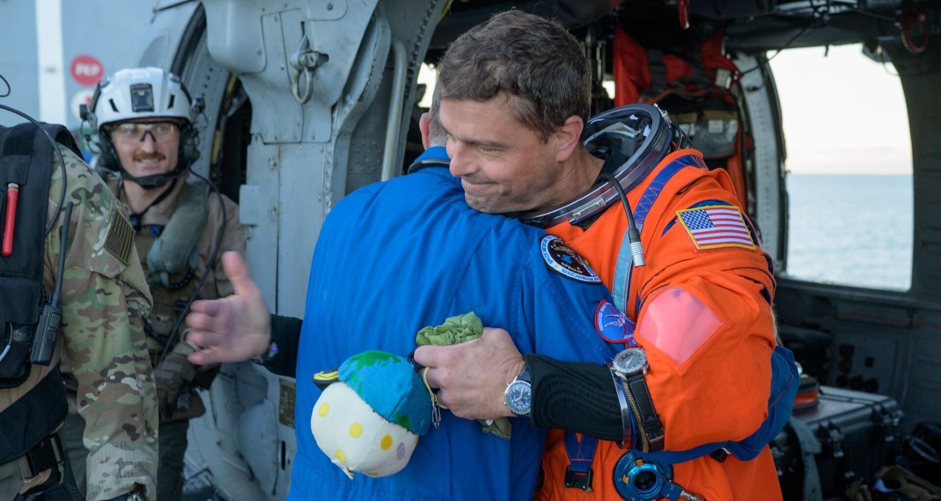 a man in an orange flight suit smiles while carrying a small stuffed moon