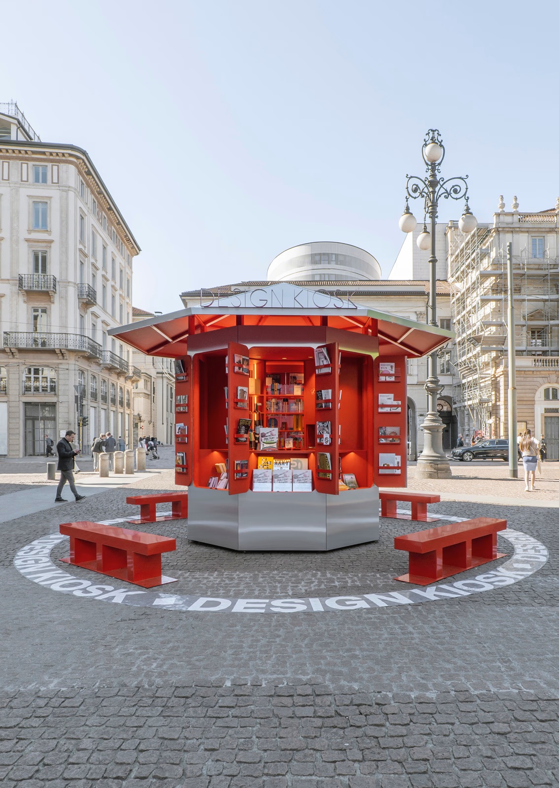 A red and chrome, retro-futuristic kiosk filled with printed matter, books, and magazines, sits in a sun-lit Italian piazza with ornate buildings.