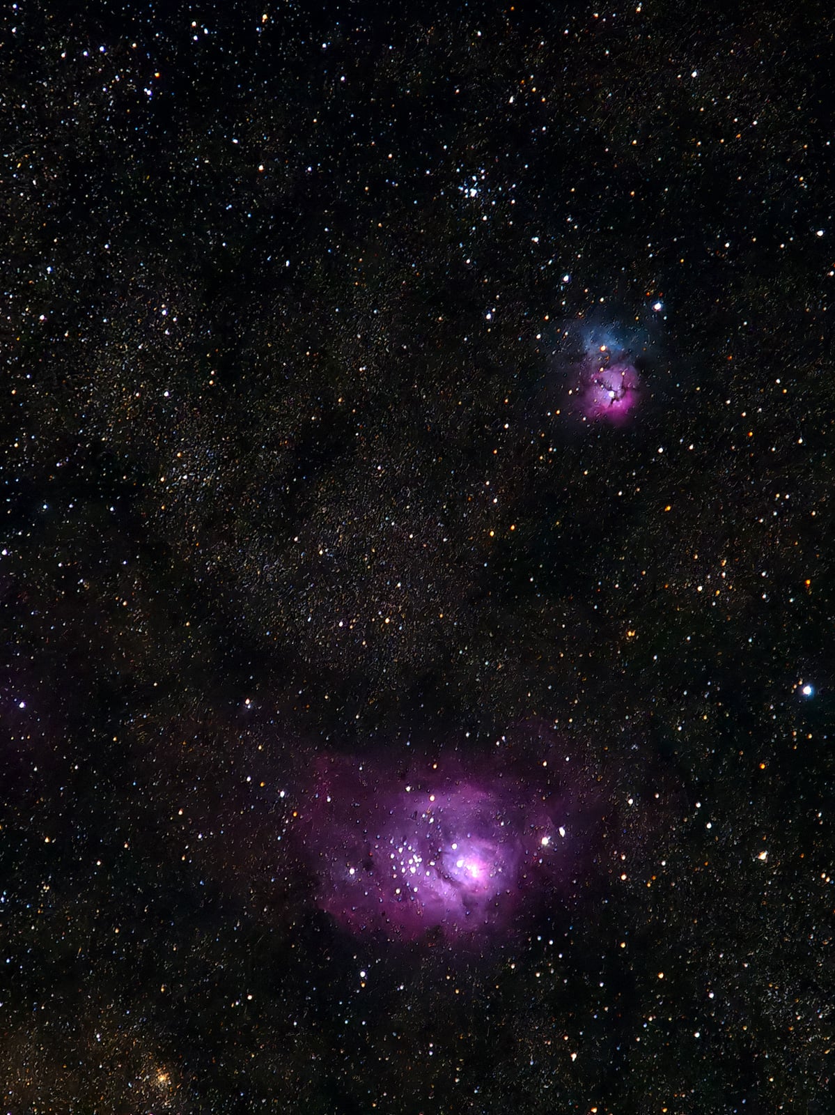 A deep-space image showing a dark star field with countless stars, featuring two bright, purple-pink nebulae—one larger nebula near the bottom and a smaller one above it—set against a black background.