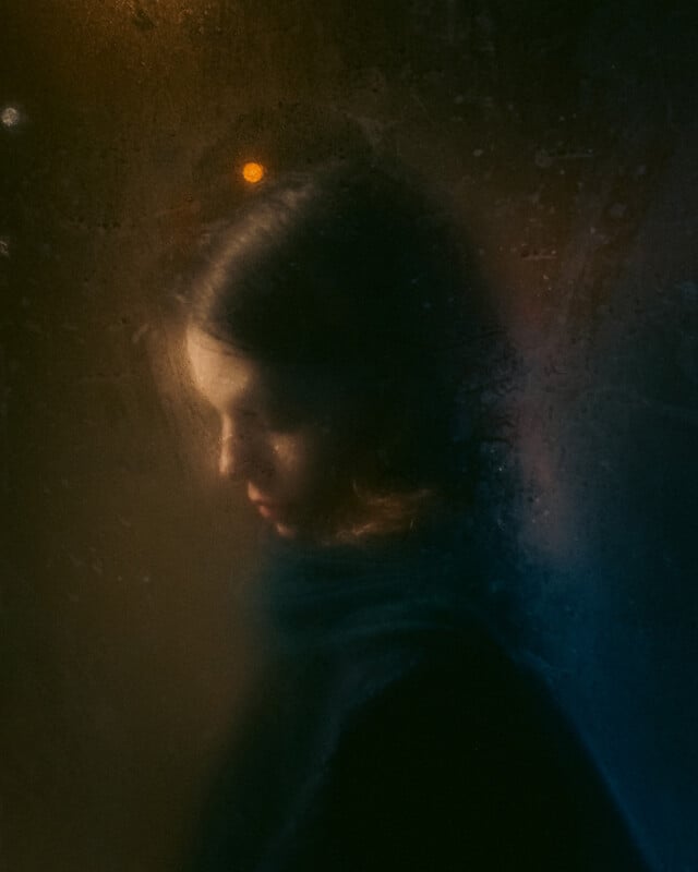 A woman with short hair is seen through a foggy, rain-streaked window. Soft, warm light illuminates her face as she looks downward, creating a moody, contemplative atmosphere.