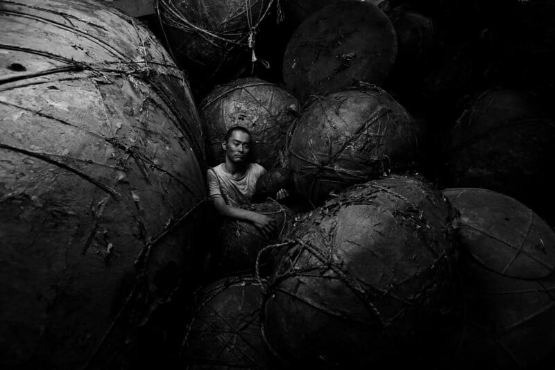 A person sits among large, round, weathered objects tied with ropes in a dimly lit, crowded space. The scene is in black and white, highlighting contrast and texture.