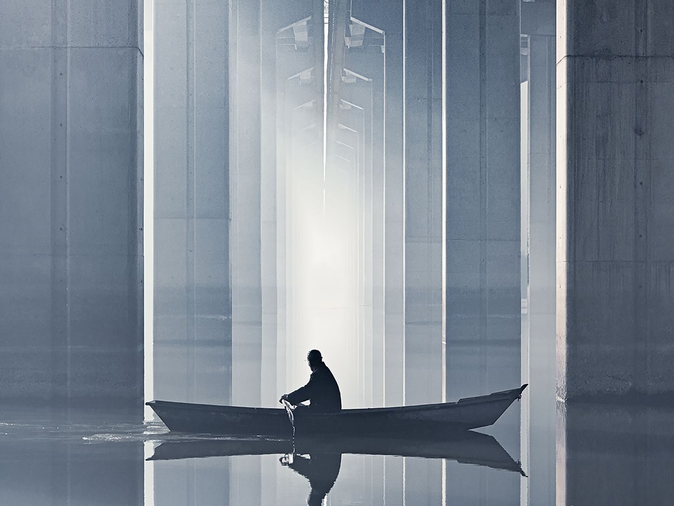 A person rows a small boat through calm waters under a modern bridge, with towering concrete beams