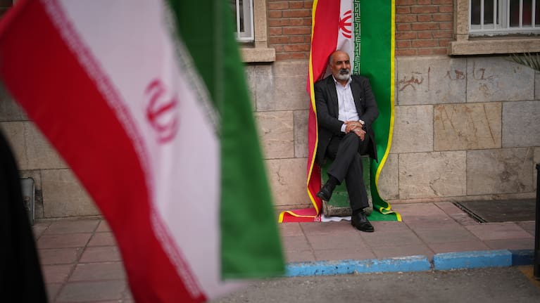 A man sits beside an Iranian flag banner during a government-sponsored protest attended by medical workers against the U.S.-Israeli military campaign outside Imam Khomeini Hospital in Tehran, Iran.