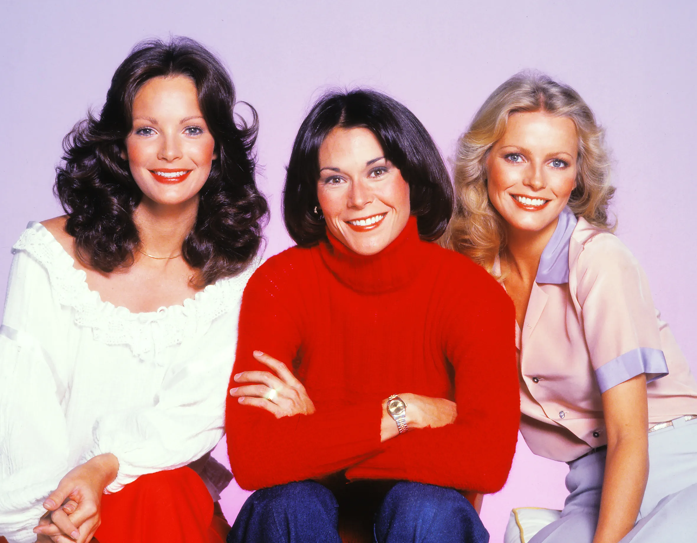 Portrait of actresses Jaclyn Smith, Kate Jackson, and Cheryl Ladd sitting on a bench against a lavender background.