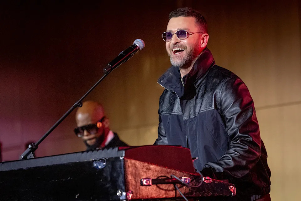 A musician in sunglasses and a leather jacket performs on stage, smiling at the audience, with a keyboard and microphone in front of them