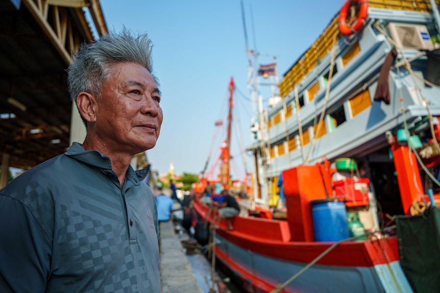 A man with wiry grey hair smiles, as he stands at a jetty next to a large multi-storey fishing boat.