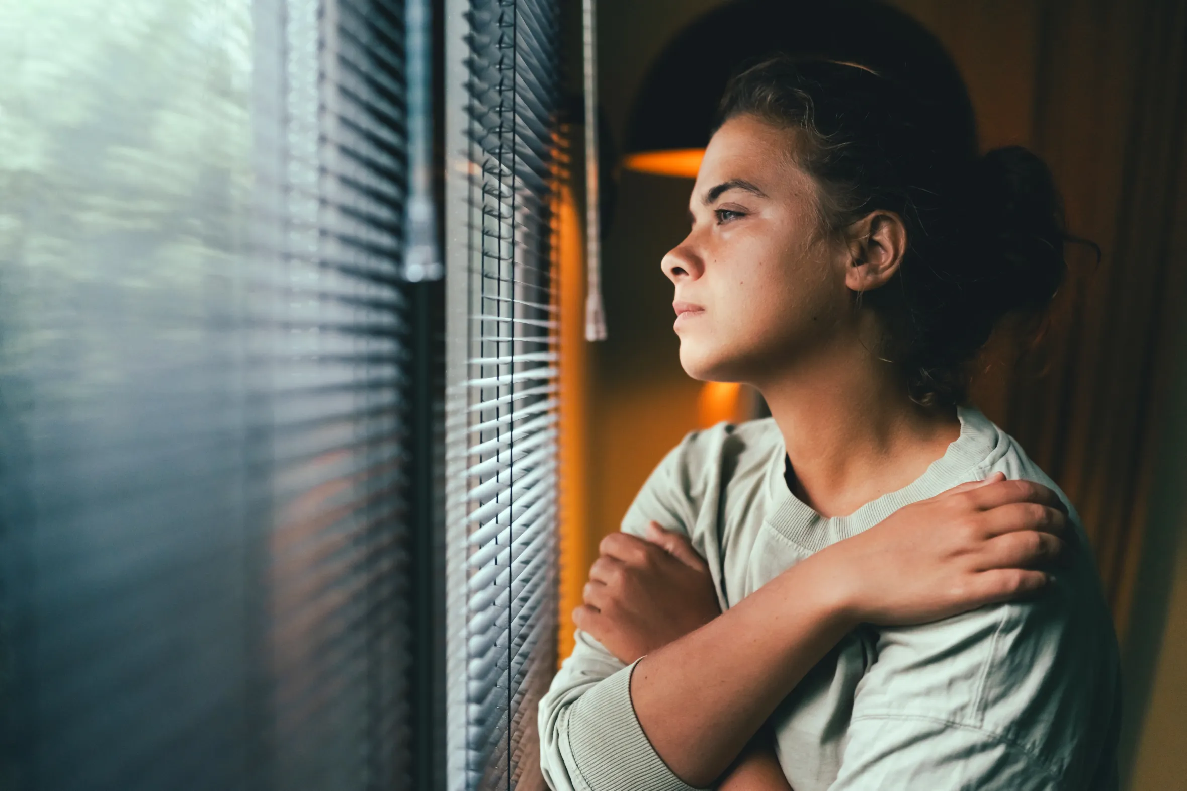 A woman with dark hair in a bun, wearing a light green shirt, looks contemplatively out a window with Venetian blinds, her arms crossed.