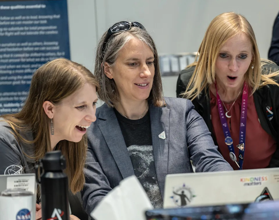 Three people excitedly look at a laptop screen, suggesting a positive discovery. They are indoors, possibly in a professional or scientific setting