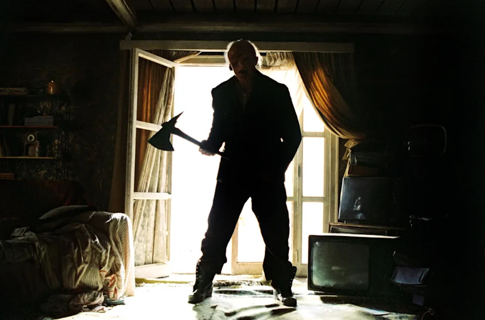 Silhouetted person holding an axe stands in front of a bright window, creating a dramatic and suspenseful atmosphere in a cluttered room